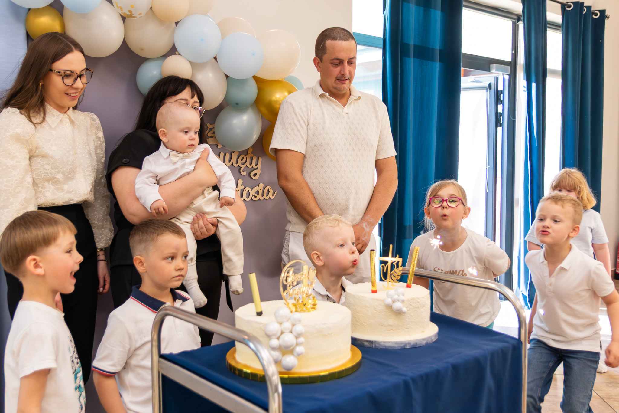 mama, chrzestni i ochrzczony oraz grupka dzieci które próbują zdmuchnąć świeczki na torcie
